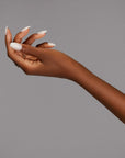 An elegant hand showcasing white almond-shaped nails with gold rings, set against a neutral gray background.