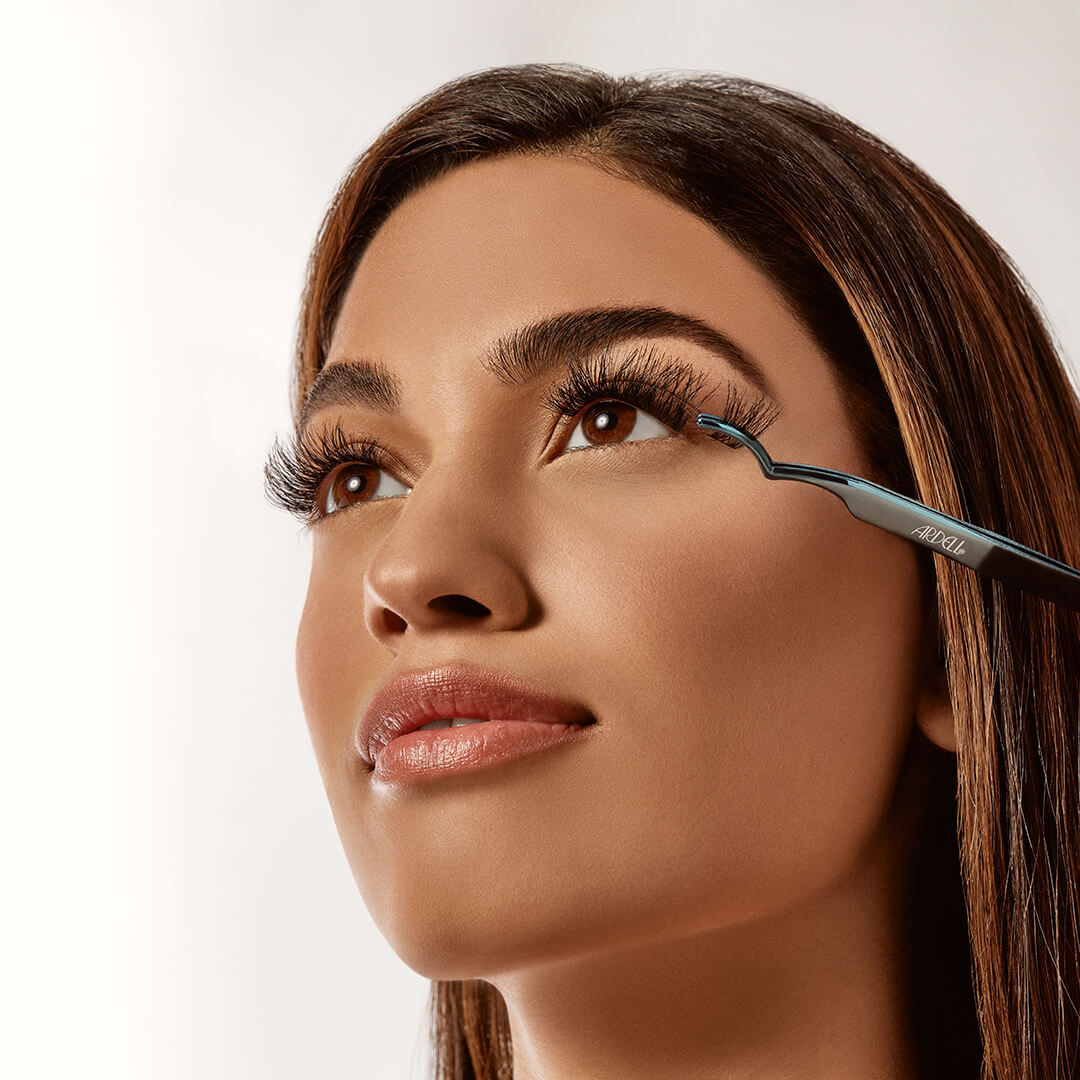 A model using Ardell tweezers to apply voluminous false eyelashes for a professional, salon-quality lash extension effect.