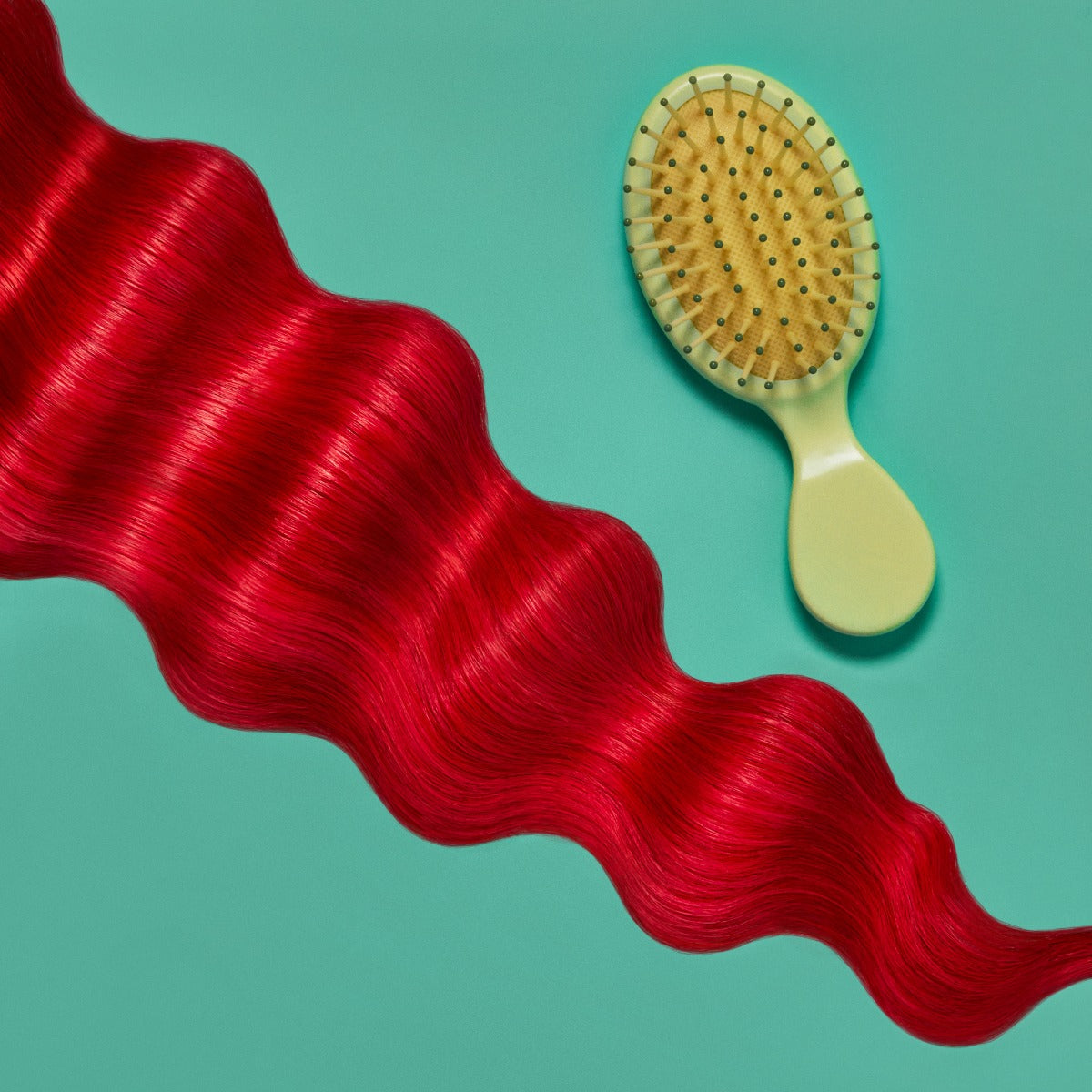 A section of wavy, bright red hair on a teal background next to a light green hairbrush, showcasing the Punky Colour Sugar Cloud Licorice Semi Permanent Foam Color