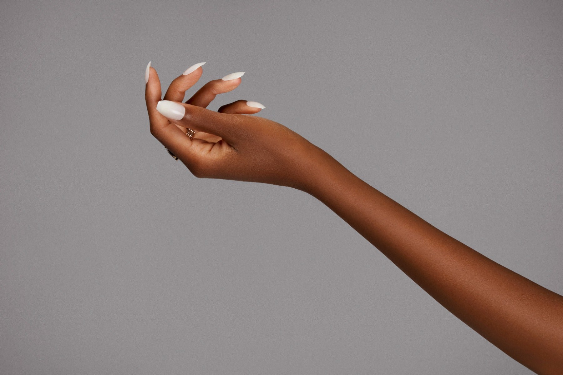 An elegant hand showcasing white almond-shaped nails with gold rings, set against a neutral gray background.
