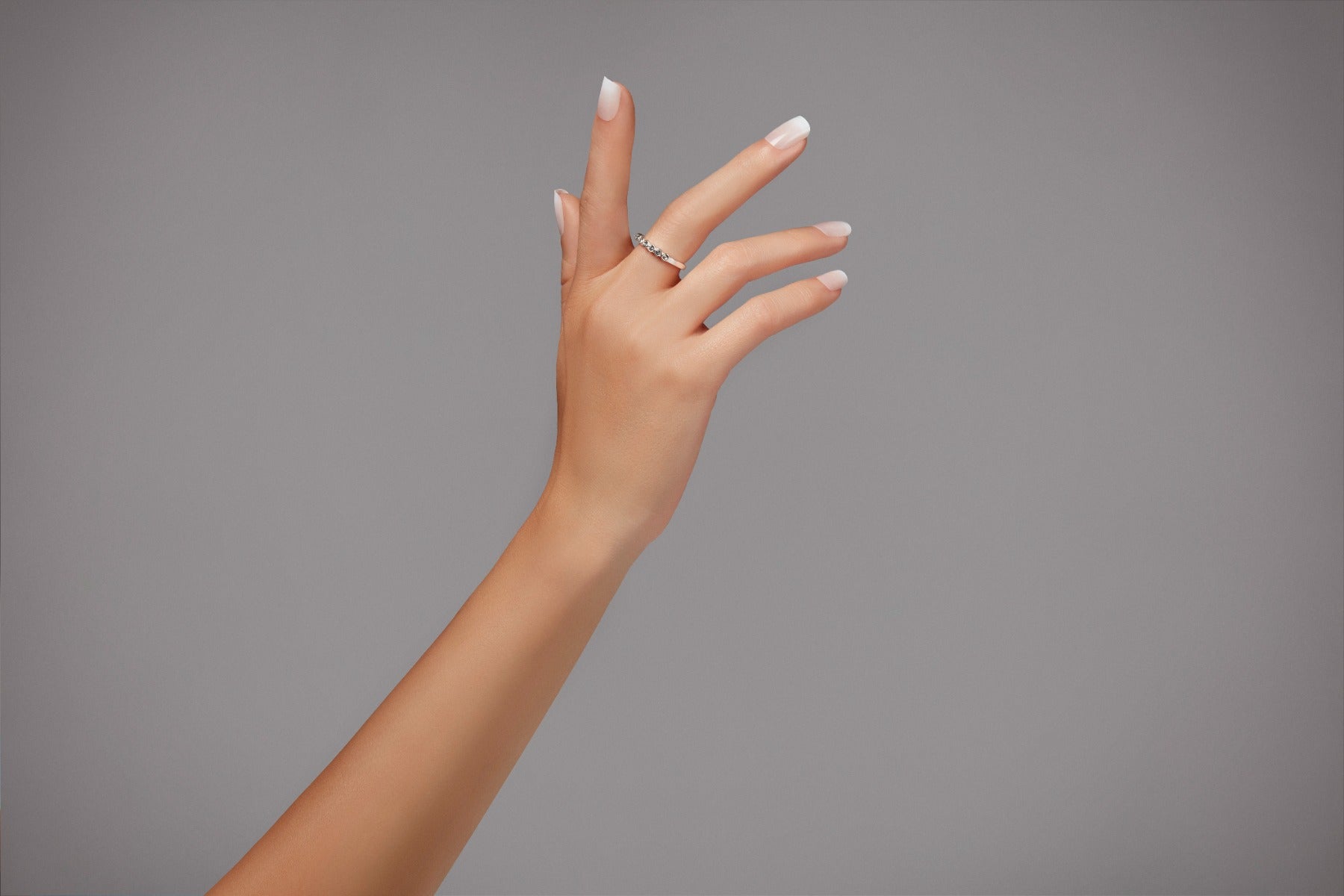 Stylish hand with natural-looking French tip nails, wearing a minimalist ring against a neutral gray background.