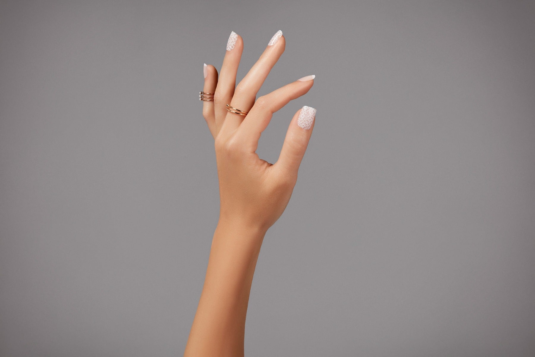 Close-up of a hand with elegant lace-patterned French tip press-on nails, complemented with gold rings.