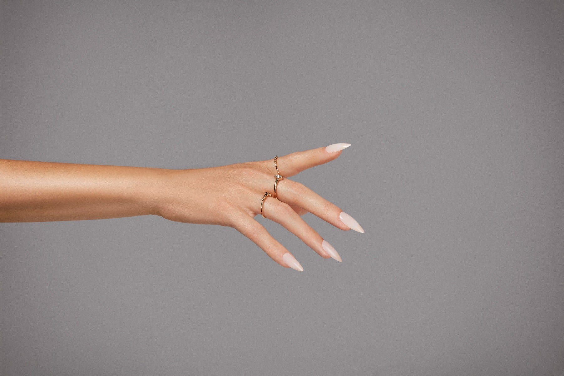 A beautifully posed hand featuring white almond-shaped nails adorned with gold rings.