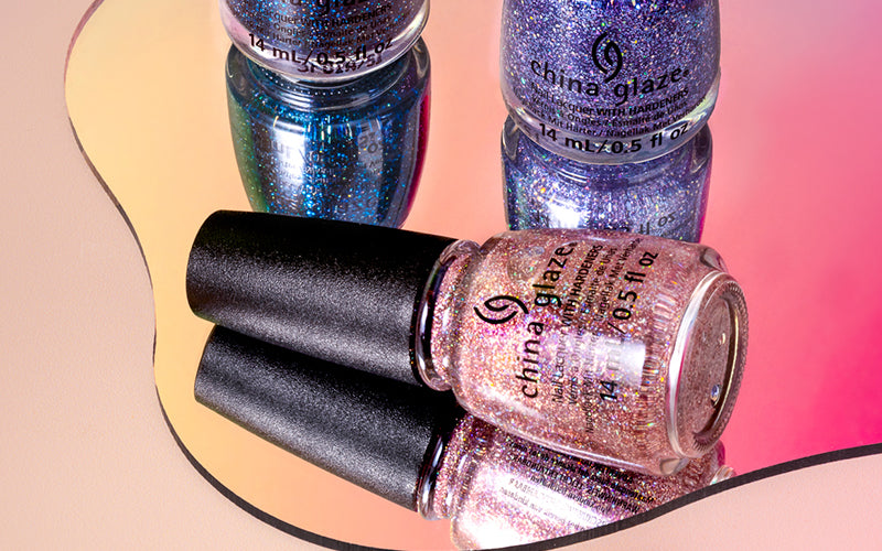 China Glaze glitter nail polish bottles in blue, purple, and pink shades with holographic shimmer reflected on a mirror over a colorful gradient background